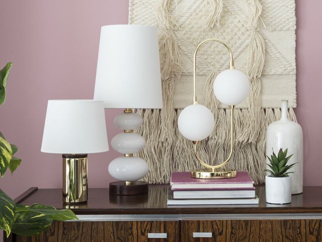 a selection of gold and brass table lamps on books on a wood sideboard with plants in front of a macrame wall hanging on a pink wall by bhs spring summer 2018