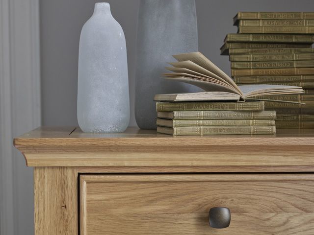 classic bedroom chest of drawers by oak furnitureland with a vase and a pile of old books on top in front of a grey wall