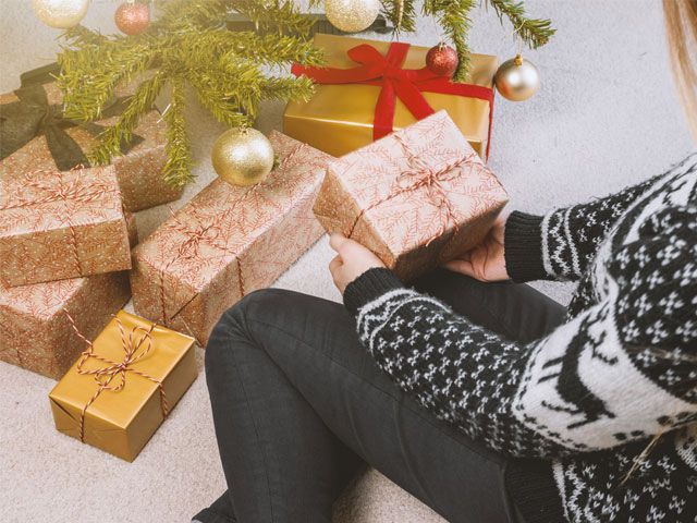 Woman wearing fair isle jumper sat holding a Christmas present underneath a Christmas tree -pexels-shopping-goodhomesmagazine.com
