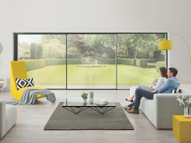 A man and woman are sat on a grey sofa in a living room with large glass panel sliding doors leading out to a garden -origin-living-room-goodhomesmagazine.com