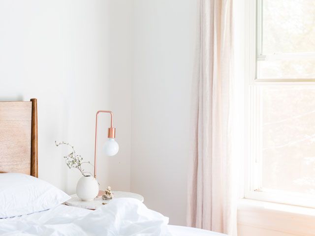 copper bedside lamp by a bed with white duvet cover and wood headboard in a bright, airy bedroom