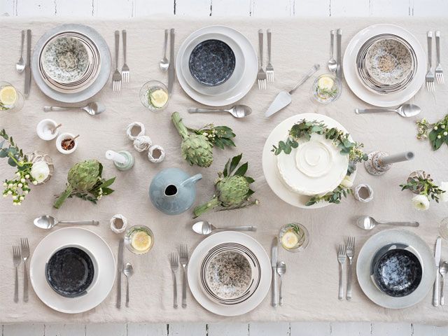 A white wooden dining table dressed with a white table cloth, green foliage and blue bowls