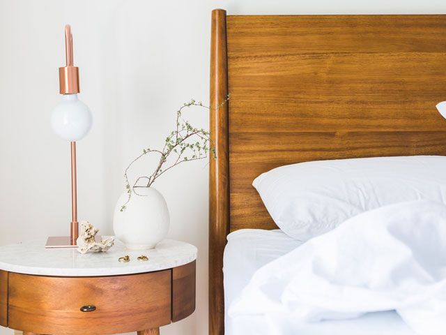 A bed with white bedding and a bedside table with earrings, a shell, copper lamp and white vase