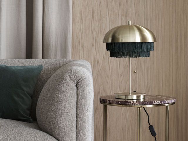 h&m home fringed brass lamp on a side table beside a grey sofa, part of H&M Home's AW19 collection