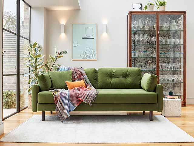 Green velvet pincushion sofa with blankets and orange scatter cushion, houseplant in background and glassware cabinet, goodhomesmagazine.com