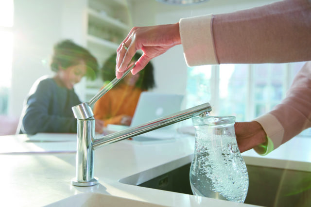 woman filling jug from kitchen tap, two people in background