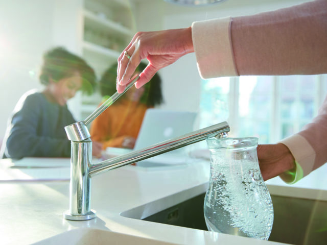 woman filling jug from kitchen tap, two people in background