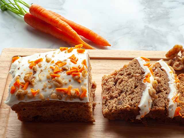 Carrot cake sliced on a wooden board with carrots in background, goodhomesmagazine.com