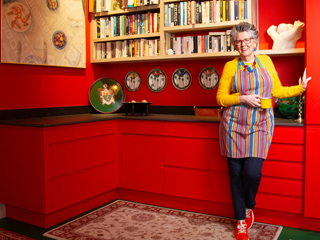 Prue Leith in her red kitchen with wall mounted plates and lots of recipe books on the shelves