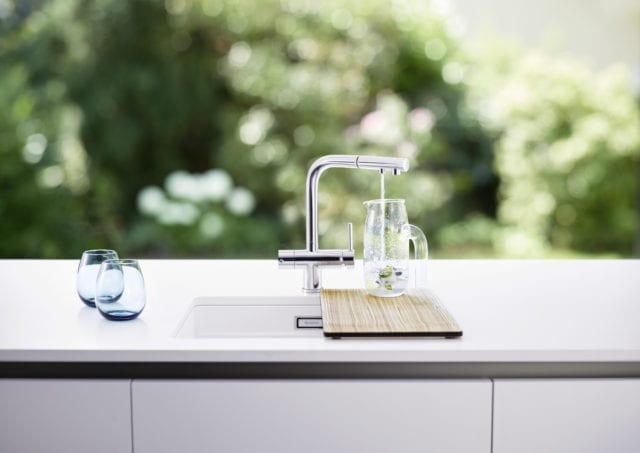 white worktop and sink with water pouring out of stainless steel tap into glass jug on wooden chopping board