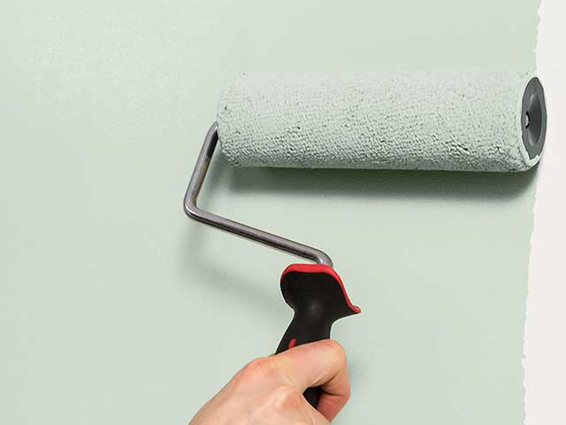 A hand using a roller brush to paint a wall mint green - Goodhomesmagazine.com