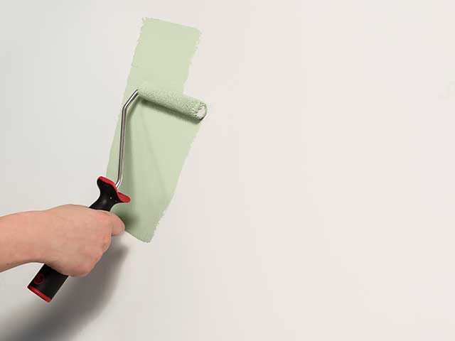 A hand using a roller brush to paint a mint green stripe on a white wall - Goodhomesmagazine.com