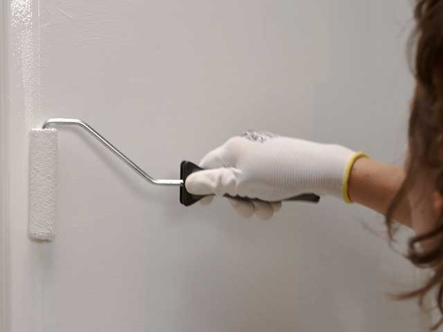 A woman using a roller brush to paint horizontally on a white wall - Goodhomesmagazine.com