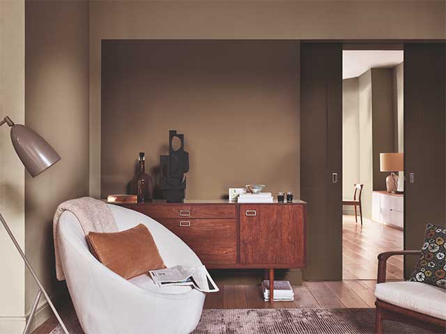 A room with brown walls, a dark wooden cabinet, and white armchair furnished with neutral accessories - Earthy tones - Goodhomesmagazine.com