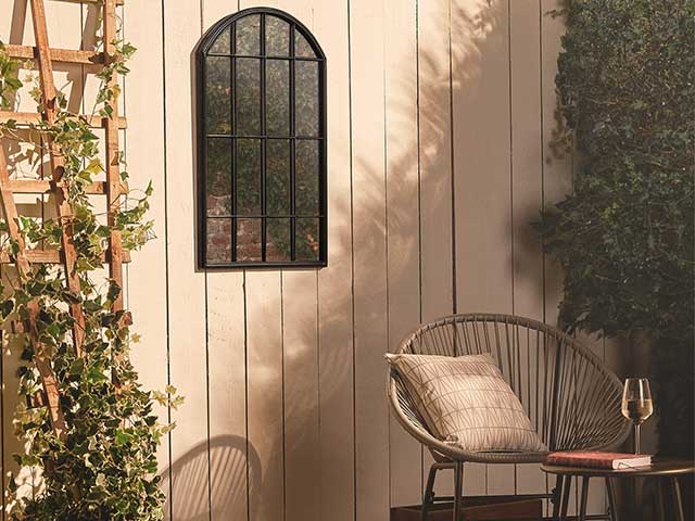 Rattan chair and plant trellis in front of a white panelled fence that has an arched mirror hanging on it - Garden mirrors - Goodhomesmagazine.com