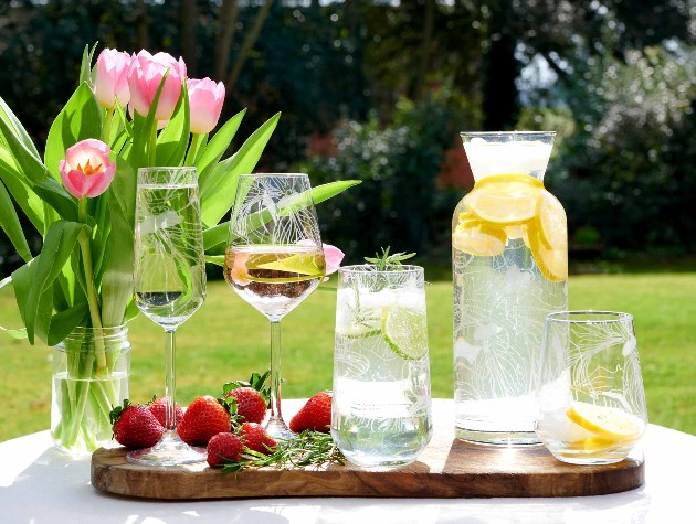 Wooden tray on a table in a summer garden with a selection of RHS glassware including champagne flute, wine glass, hi ball and carafe etched with iris design