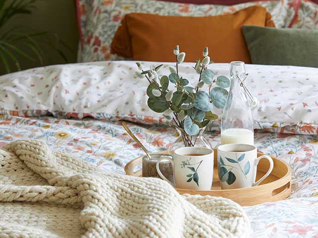 A bed with heritage pattern bedding, a wooden tray with mugs on it, a woven throw, and a burnt orange cushion - Goodhomesmagazine.com