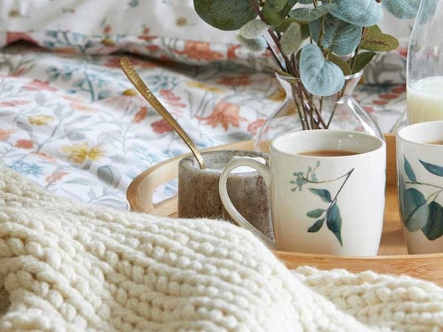 A bed with heritage pattern bedding, a wooden tray with mugs on it, a woven throw, and a burnt orange cushion - Goodhomesmagazine.com