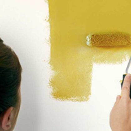 Woman using a roller brush and yellow paint to paint an area on the wall that has been marked out with masking tape - Painted headboards - Goodhomesmagazine.com