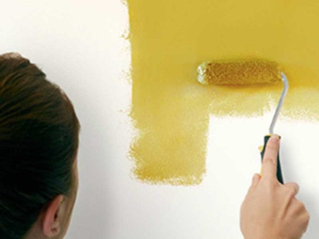 Woman using a roller brush and yellow paint to paint an area on the wall that has been marked out with masking tape - Painted headboards - Goodhomesmagazine.com