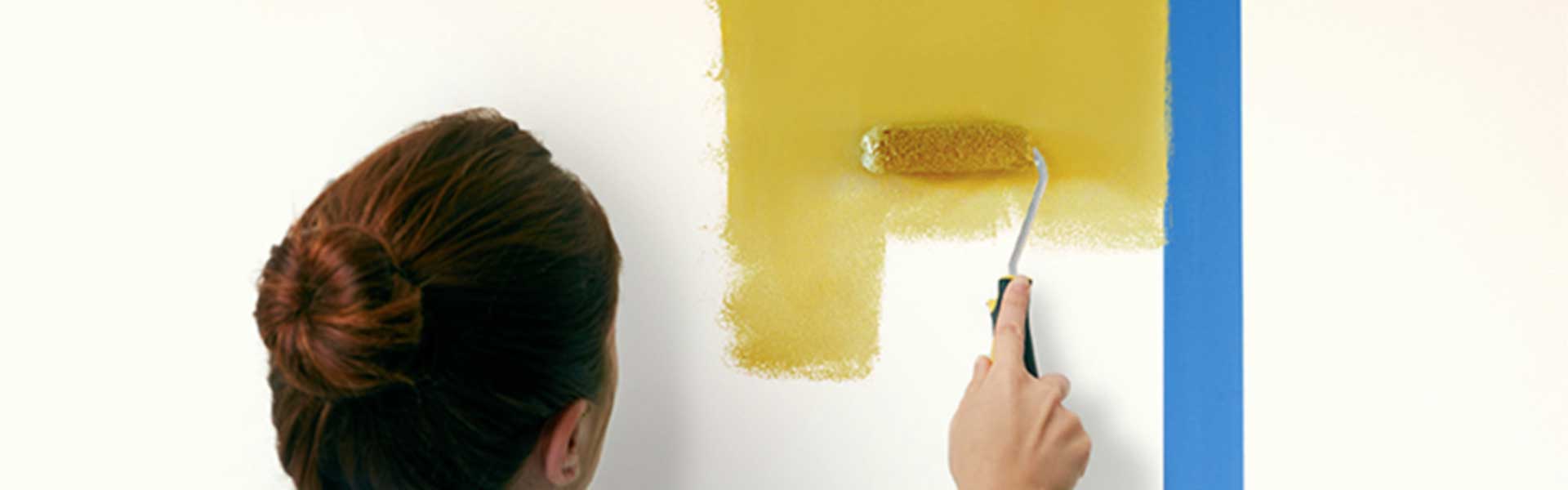 Woman using a roller brush and yellow paint to paint an area on the wall that has been marked out with masking tape - Painted headboards - Goodhomesmagazine.com