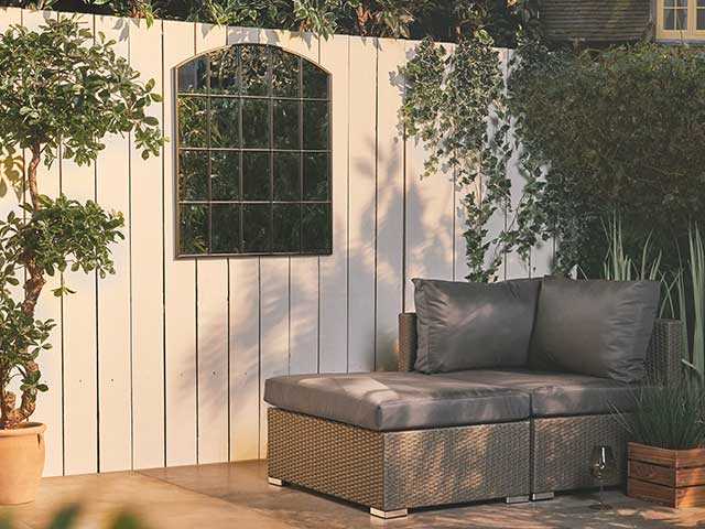 Grey rattan sofa in front of a white panelled fence with a gothic mirror hanging on it - Garden mirrors - Goodhomesmagazine.com