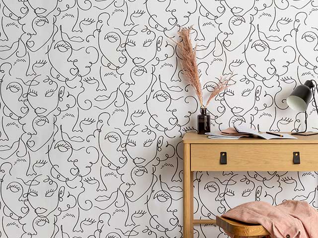 Wooden desk with pink accessories in front of a wall featuring wallpaper with abstract faces - Goodhomesmagazine.com