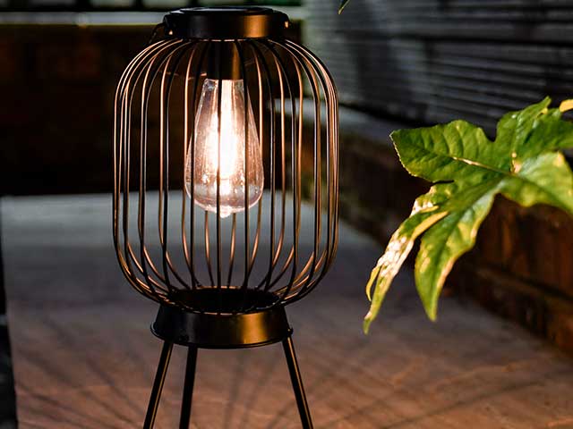 Copper coloured caged lantern on three metal legs on garden pathway with foilage in background, goodhomesmagazine.com