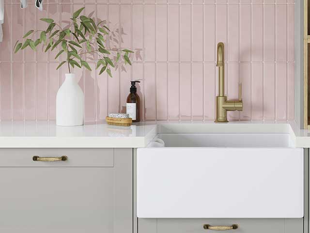 A deep white Belfast sink with a gold tap in front of a pink tiled backsplash
