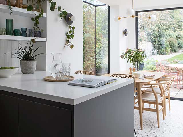Open plan kitchen in this Melbourne inspired home with dark units and white worktops and dining table in background with floor length windows