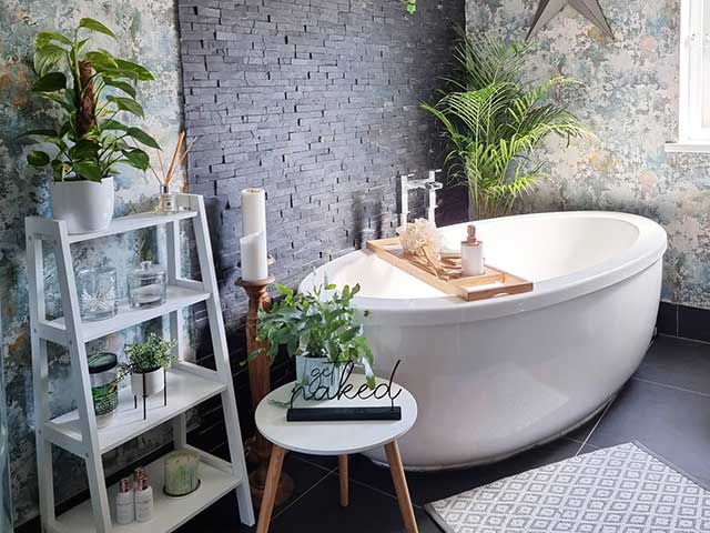 Freestanding bath in plant-filled bathroom with black tiled wall and black flooring, white shelving unit and plenty of houseplants