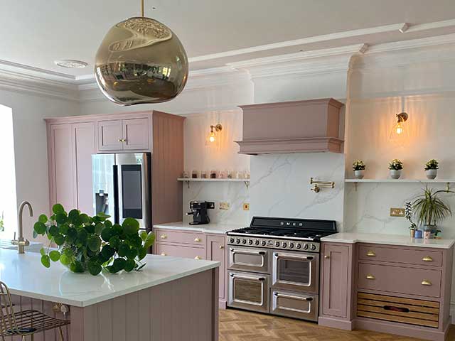 Pink units in kitchen with wooden flooring and pink kitchen island with gold taps and handles, and gold light fitting