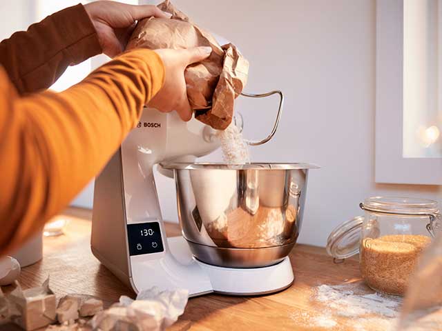 Person pouring flour into kitchen mixer machine on wooden worktop with glass jars in background