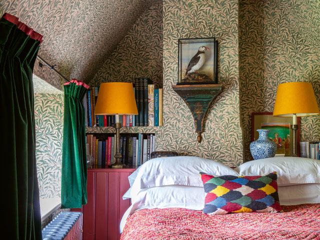 A cosy bedroom in a countryside cottage with leaf-print wallpaper and colourful bedspread and curtains