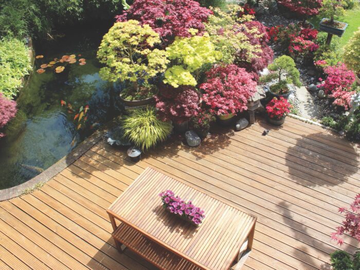 Photo showing a landscaped contemporary Japanese garden with a large expanse of pre-treated timber decking, providing a family space for outdoor furniture - a modern slatted table and benches. The stained decking has been cut to form a curve around a large koi pond, while other features include Japanese maples, bonsai trees and oriental lanterns.